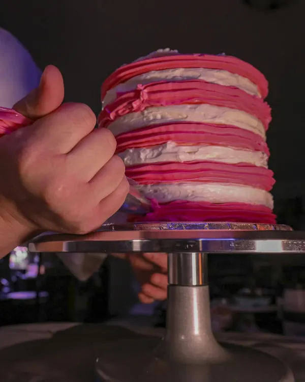 À l’atelier, une main poche de la crème rose sur un gâteau posé sur une tournette